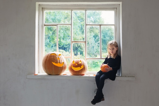 Kleine heks in zwarte jurk met halloween pompoenen op vensterbank