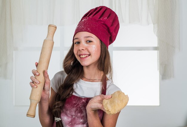 Kleine helper rolt het deeg met deegroller culinair en keuken gelukkige jeugd gelukkig kind slijtage kok uniform chef-kok meisje in hoed en schort kind koken voedsel in de keuken een carrière kiezen