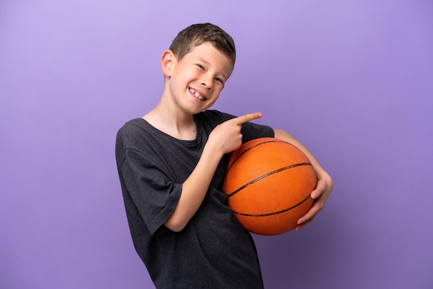 Kleine jongen die basketbal speelt geïsoleerd op een paarse achtergrond die met de vinger naar de zijkant wijst