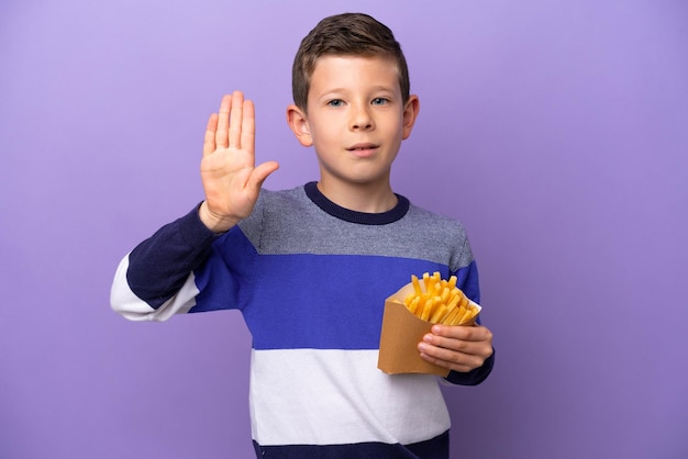 Kleine jongen die gefrituurde chips vasthoudt die op een paarse achtergrond worden geïsoleerd en een stopgebaar maakt