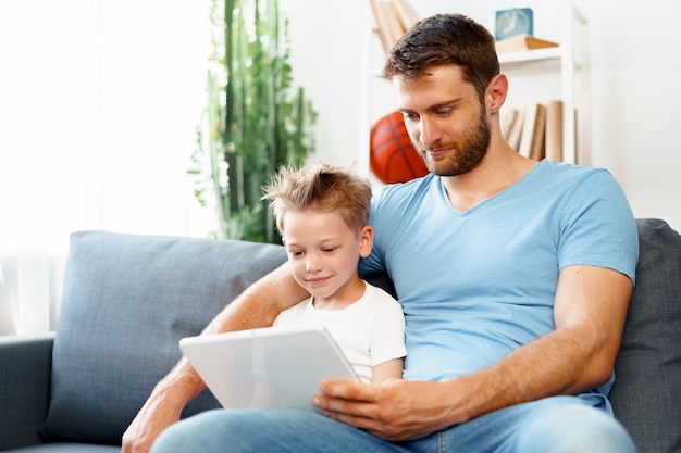 Kleine jongen en zijn vader kijken naar video's op digitale tablet samen zittend op de bank thuis