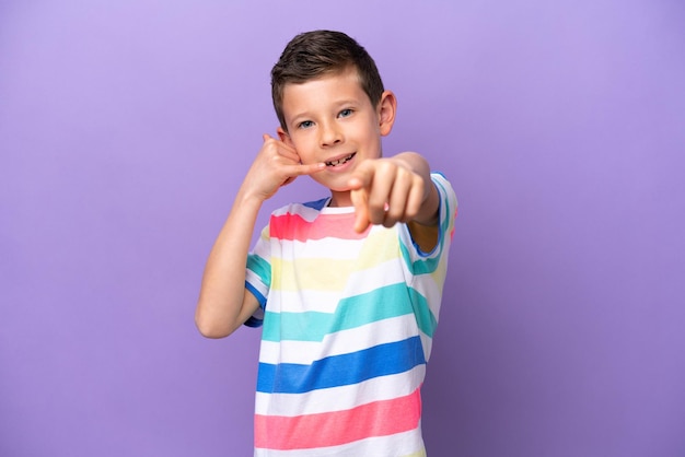 Kleine jongen geïsoleerd op een paarse achtergrond die een telefoongebaar maakt en naar voren wijst