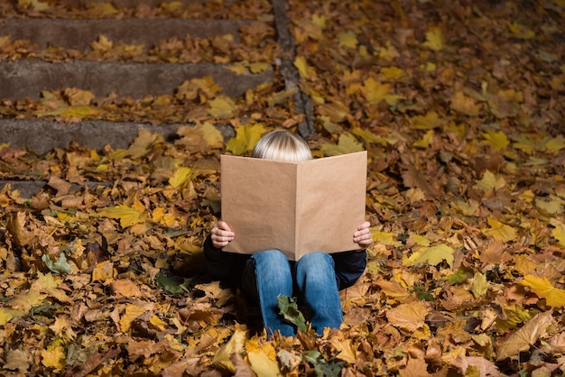 Kleine jongen groot boek in handen houden en zittend op gevallen herfstbladeren. Kind houdt van lezen.