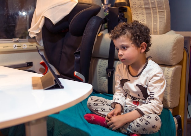 Kleine jongen in camper vermaakt zichzelf door naar tablet te kijken terwijl hij in pyjama zit