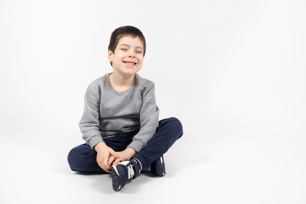 Kleine jongen in een grijs sweatshirt, blauwe trainingsbroek en sneakers op een witte achtergrond zit en glimlacht ruimte voor tekst