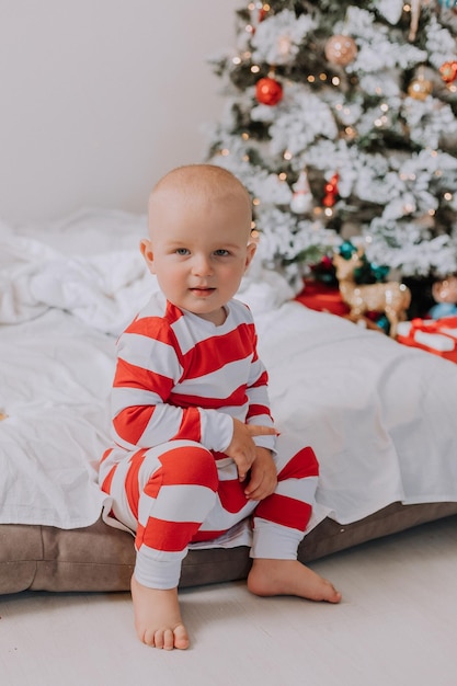 Kleine jongen in rood-witte pyjama zit op de rand van het bed naast de kerstboom