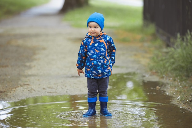 Kleine jongen in waterdichte blauwe rubberen laarzen in plassen met plons na de regen