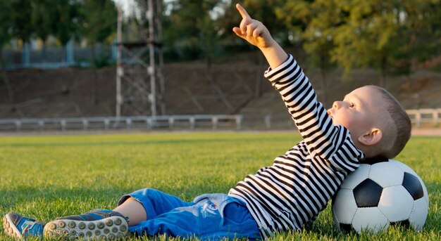 Kleine jongen liggend op zijn rug in het groene gras met zijn hoofd rustend op een voetbal die naar de hemel wijst