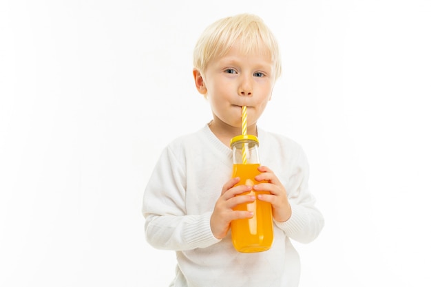 Kleine jongen met kort blond haar, blauwe ogen, schattig uiterlijk, in witte jas, lichtblauwe broek, stands en sinaasappelsap drinkt uit een glazen fles met een gestreepte buis