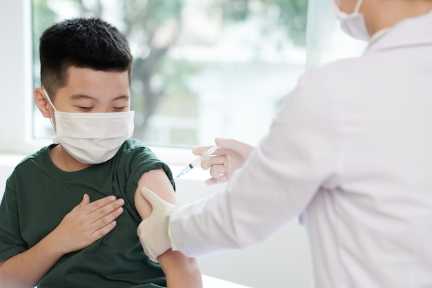 Kleine jongen met medisch masker laat dokter vaccin injecteren tegen coronavirus