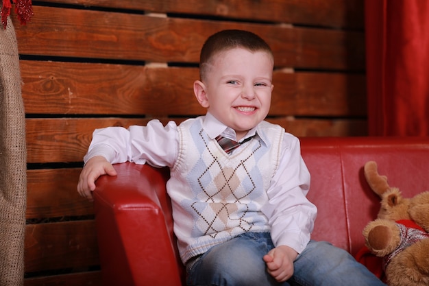 Foto kleine jongen met plezier op de bank in de kerststudio in het rood. kerstboom en teddybeer op de muur.