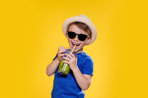 Kleine jongen met strohoed en zonnebril die mojito drinkt in plastic beker op gele achtergrond