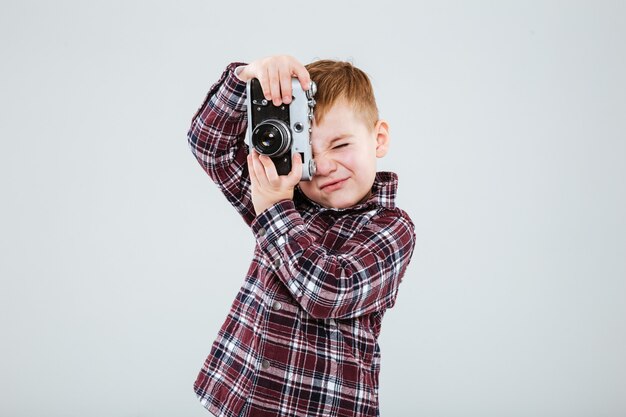 Kleine jongen staat en maakt foto's met oude vintage fotocamera over witte muur