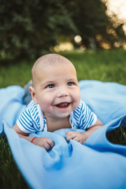 Kleine jongen van 3 maanden oud ligt op zijn buik op een blauwe sprei in het park rond het groene gras en de bomen. Emoties van vreugde bij kinderen