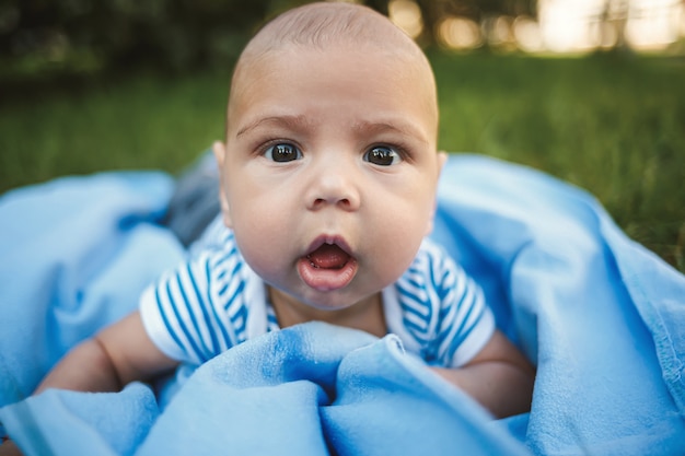 Kleine jongen van 3 maanden oud ligt op zijn buik op een blauwe sprei in het park rond het groene gras en de bomen. Emoties van vreugde bij kinderen