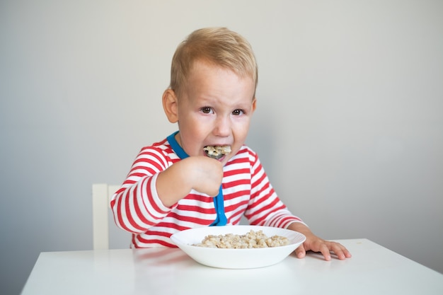 kleine jongen zit aan tafel en eet havermout met een grote lepel