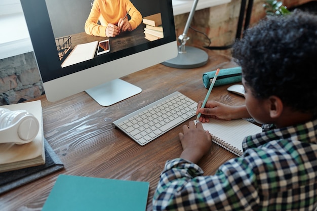 Kleine jongen zit aan tafel voor computermonitor tijdens online les, luistert naar zijn leraar op het scherm en maakt aantekeningen in notitieboekje