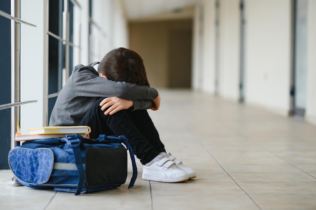 Kleine jongen zit alleen op de vloer nadat hij gepest is terwijl kinderen op de achtergrond rennen Trieste jonge schooljongen zittend op de gang met handen op knieën en hoofd tussen zijn benen