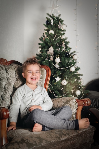 Kleine jongen zit bij de kerstboom. Gelukkig kind, kerstviering.