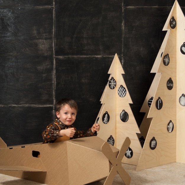 Kleine jongen zit in een kartonnen speelgoedvliegtuigje in de buurt van houten kerstbomen