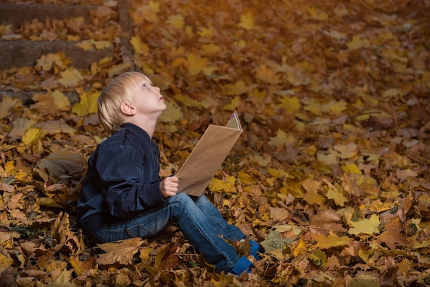 Kleine jongen zit in een wild bos in herfstbladeren met een boek in handen Kinderboeken over wonderen en magie