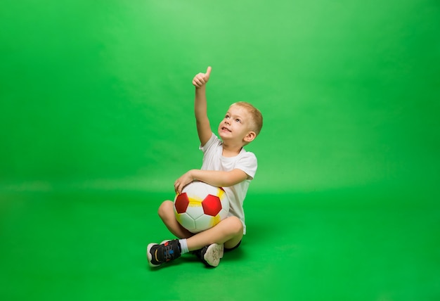 Kleine jongen zit in uniform en met een voetbal en toont zijn hand aan de klas op groen