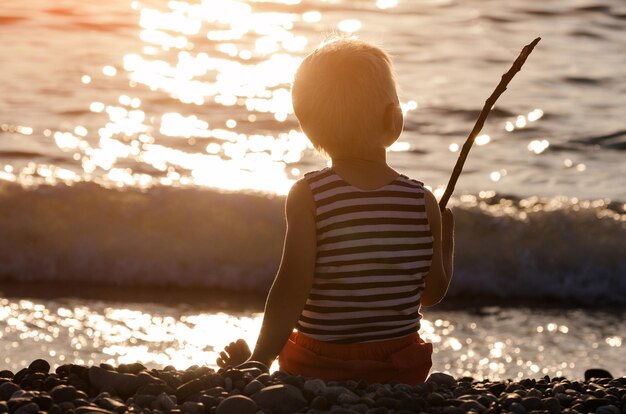 Kleine jongen zit op het strand bij zonsondergang en houdt een geïmproviseerde hengel vast