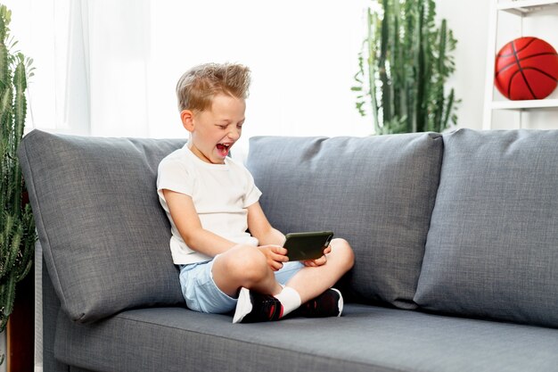 Kleine jongen zittend op de bank thuis en met behulp van smartphone