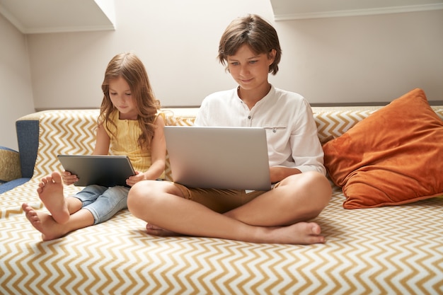 Kleine kinderen schattige broer en zus zittend op de bank en met behulp van laptop en digitale tablet