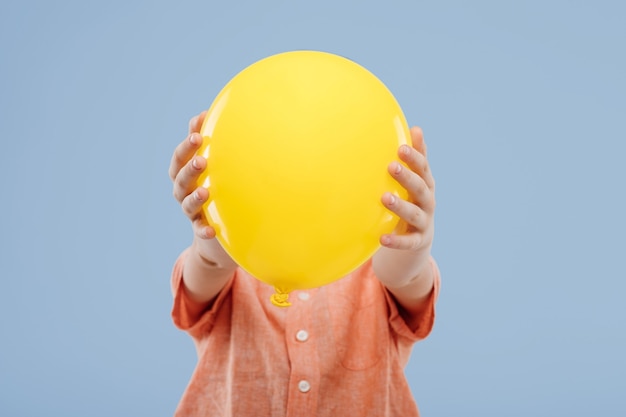 Kleine kindjongen die gezicht bedekt met gele ballon geïsoleerd op blauwe achtergrond kopie ruimte