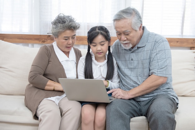 Kleine kleindochter leert senior oudere om te surfen op internet met behulp van computer en technologie en moderne levensstijl. Gelukkige Aziatische grootouder met kleine jonge schattige kleinkind zittend op de bank spelen laptop om
