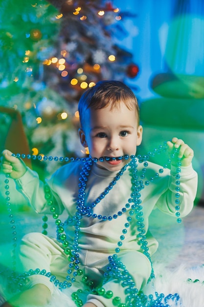 Foto kleine knappe eenjarige jongen gekleed in een beige pak op de achtergrond van een kerstboom in een thuisomgeving kinderemoties voordat hij de nieuwjaarsvakantie viert