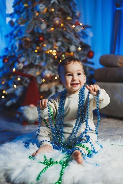 Kleine knappe eenjarige jongen gekleed in een beige pak op de achtergrond van een kerstboom in een thuisomgeving Kinderemoties voordat hij de nieuwjaarsvakantie viert