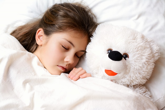 Kleine mooie meid met lang donker haar gezonde huid ligt op comfortabel bed met haar favoriete speeltje. Meisjeslaap met haar teddybeer onder de witte warme deken heeft prettige dromen 's nachts