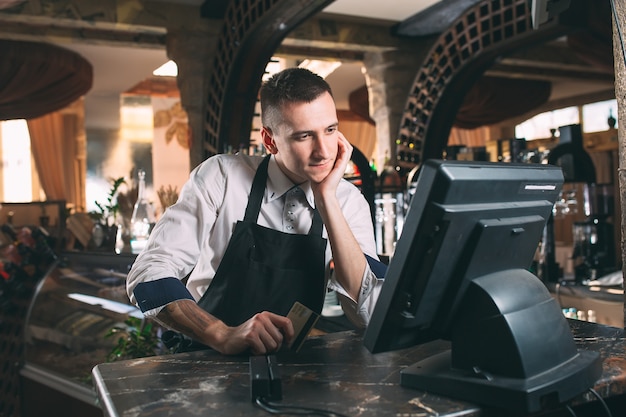 Kleine onderneming, mensen en de dienstconcept - gelukkige mens of kelner in schort bij teller met kassa werken bij bar of koffiewinkel