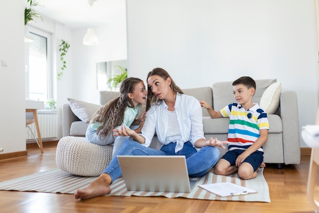 Kleine ondeugende kinderen die moeder freelancer lawaai maken en afleiden die zich op laptop probeert te concentreren en hoofd samenknijpt aan tafel in lichte woonkamer