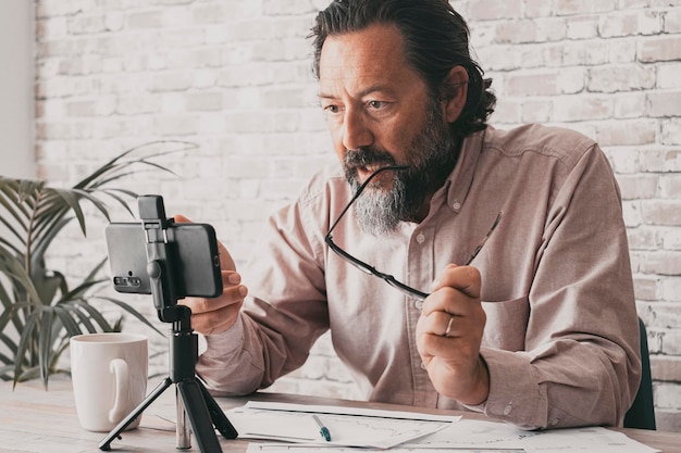 Kleine online zakenman praten in videogesprek voor een mobiele telefoon op statief in kantoor thuiswerkplek Papieren documenten op bureau Professioneel freelance werken digitaal met mobiele communicatie