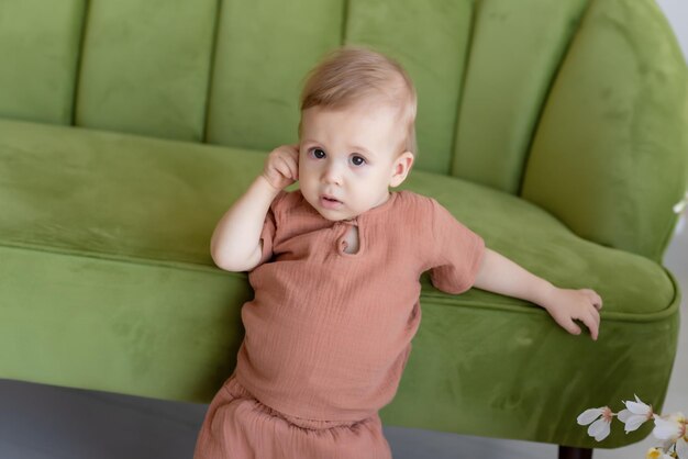 Kleine pasgeboren jongen in een beige pak staat in de buurt van een groene bank omringd door prachtige bloemen Kijkend naar de camera