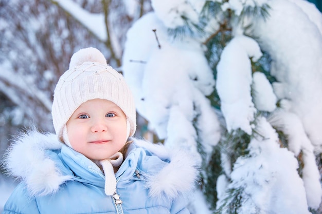 Kleine peuterjongen in blauwe jas en gebreide muts die op een zonnige dag in een besneeuwd bos of park staat, smailend en naar de camera kijkt.