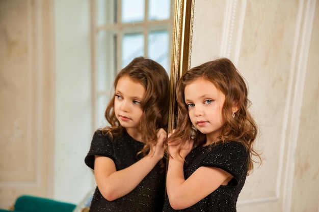 Foto kleine prinses 67 jaar oud blond meisje model met blauwe ogen in stijlvolle zwarte elegante jurk in de spiegel in de woonkamer kijkend naar de camera