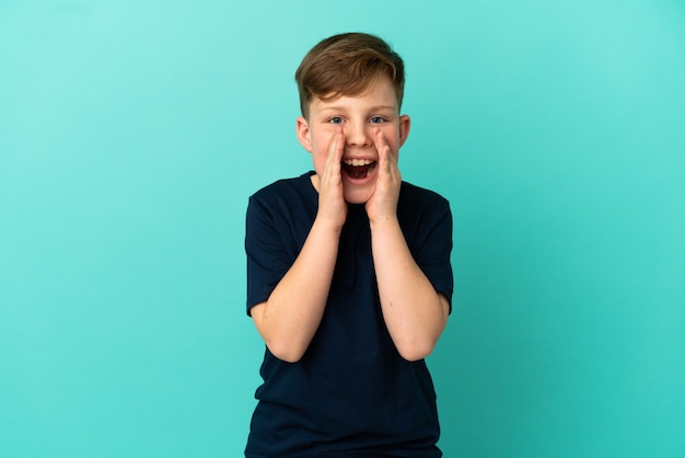 Kleine roodharige jongen geïsoleerd op blauw oppervlak schreeuwen en iets aankondigen