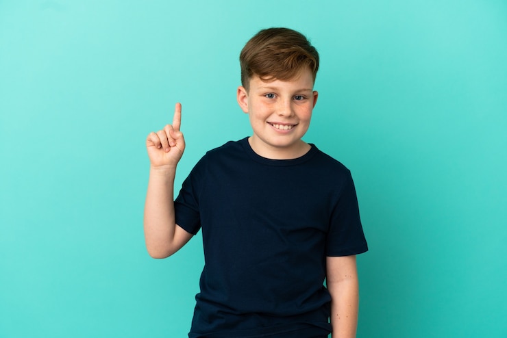 Foto kleine roodharige jongen geïsoleerd op een blauwe muur die een vinger toont en optilt in teken van de beste