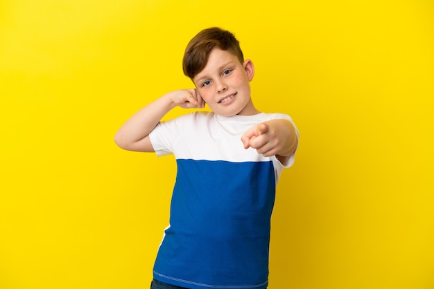 Kleine roodharige jongen geïsoleerd op een gele achtergrond die een telefoongebaar maakt en naar voren wijst