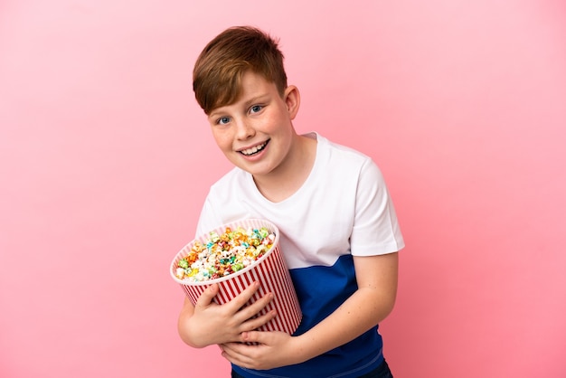 Kleine roodharige jongen geïsoleerd op roze achtergrond met een grote emmer popcorns