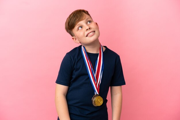 Kleine roodharige jongen met medailles geïsoleerd op roze achtergrond terwijl hij glimlacht