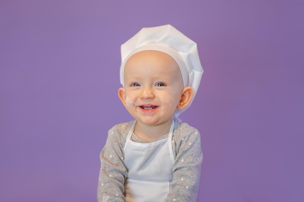 Kleine schattige baby in een schort en een koksmuts glimlacht op een lila achtergrond geïsoleerd