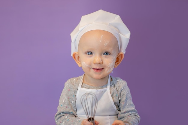 Kleine schattige baby in een schort en een koksmuts glimlacht op een lila achtergrond