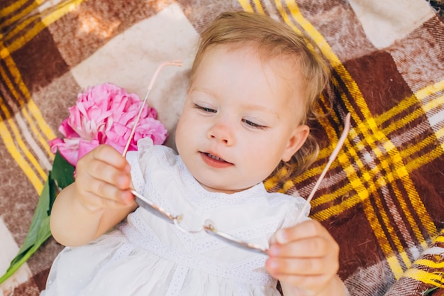 Kleine schattige baby in een witte jurk met een roze pioen in handen liggen op een plaid in het park. Familie openluchtrecreatie. Familie zomerpicknick in het park. Meisje buiten omgeven door roze pioenrozen.