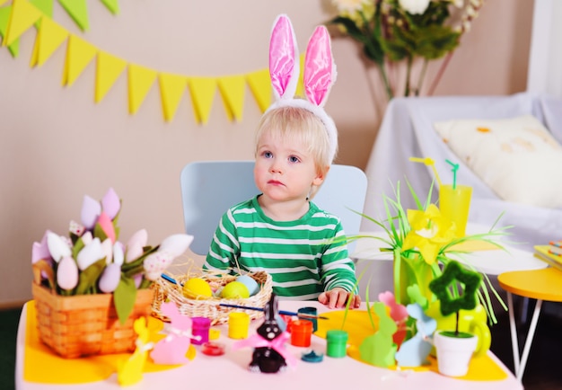 Kleine schattige babyjongen met wit haar met konijnenoren op zijn hoofd zitten aan een tafel voor een mand en decor van Pasen.