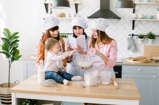 Kleine schattige babymeisje helpt haar moeder, tante en grootmoeder om samen een deeg te maken. grootmoeder leest recept van koekjes uit het boek. gelukkige familie bakken in de keuken